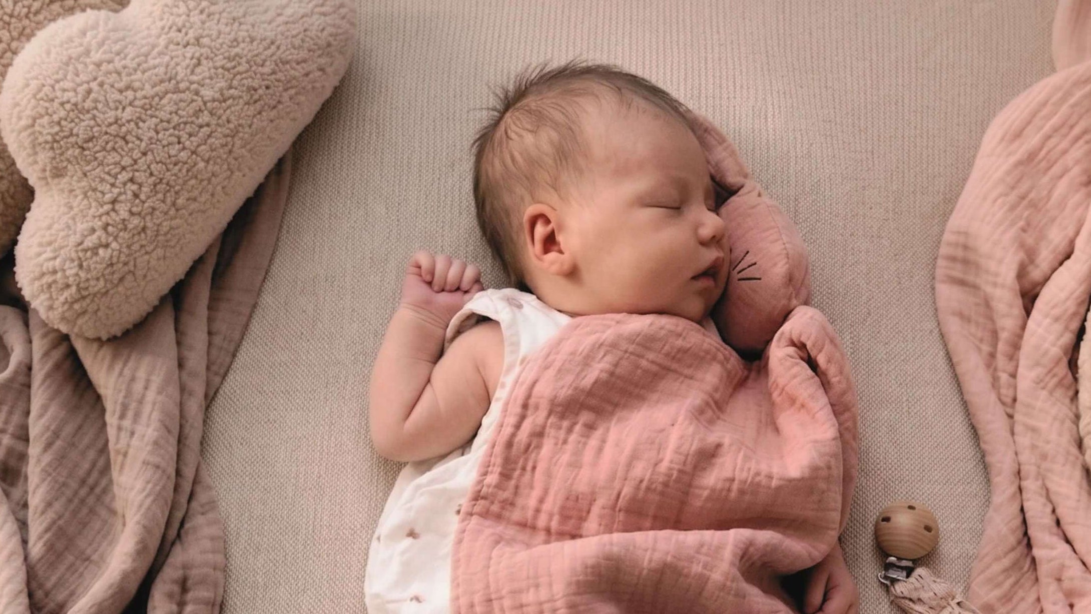 Friedlich schlafendes Baby in weißem Musselin-Body auf beiger Strickdecke, skandinavisches Kinderzimmer-Design mit altrosa Musselin-Tuch und mondförmigem Kissen